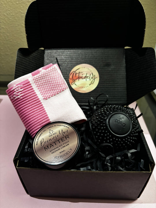 A gift box set from Stridely featuring a pink and white striped knit scarf, a tin of Becca's Art MATTER Advanced Hair Removal Cream, and a black textured silicone scrubber, all arranged in a black gift box with black paper shred.