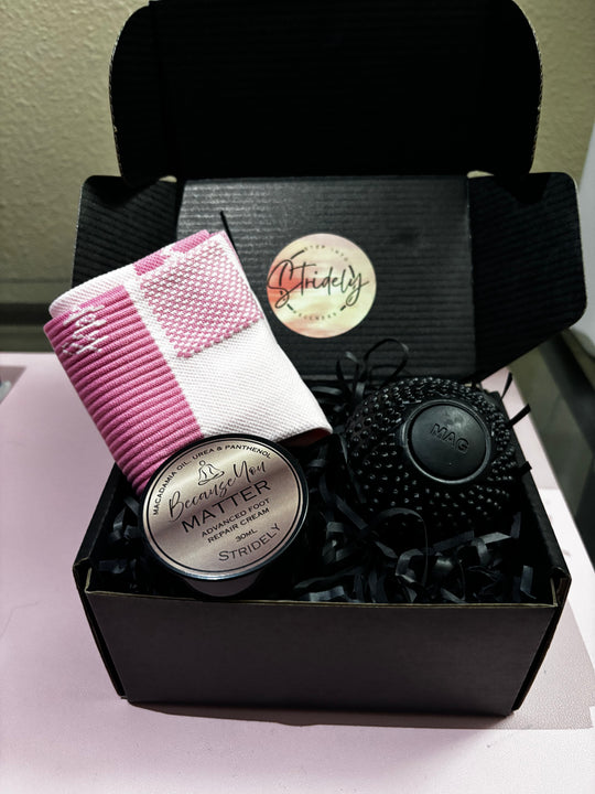 A black gift box filled with beauty products, including a pink and white striped silk scarf, a round tin labeled 'Beauty Matter' by Sydney, and a black textured round item, all arranged in black paper shred.